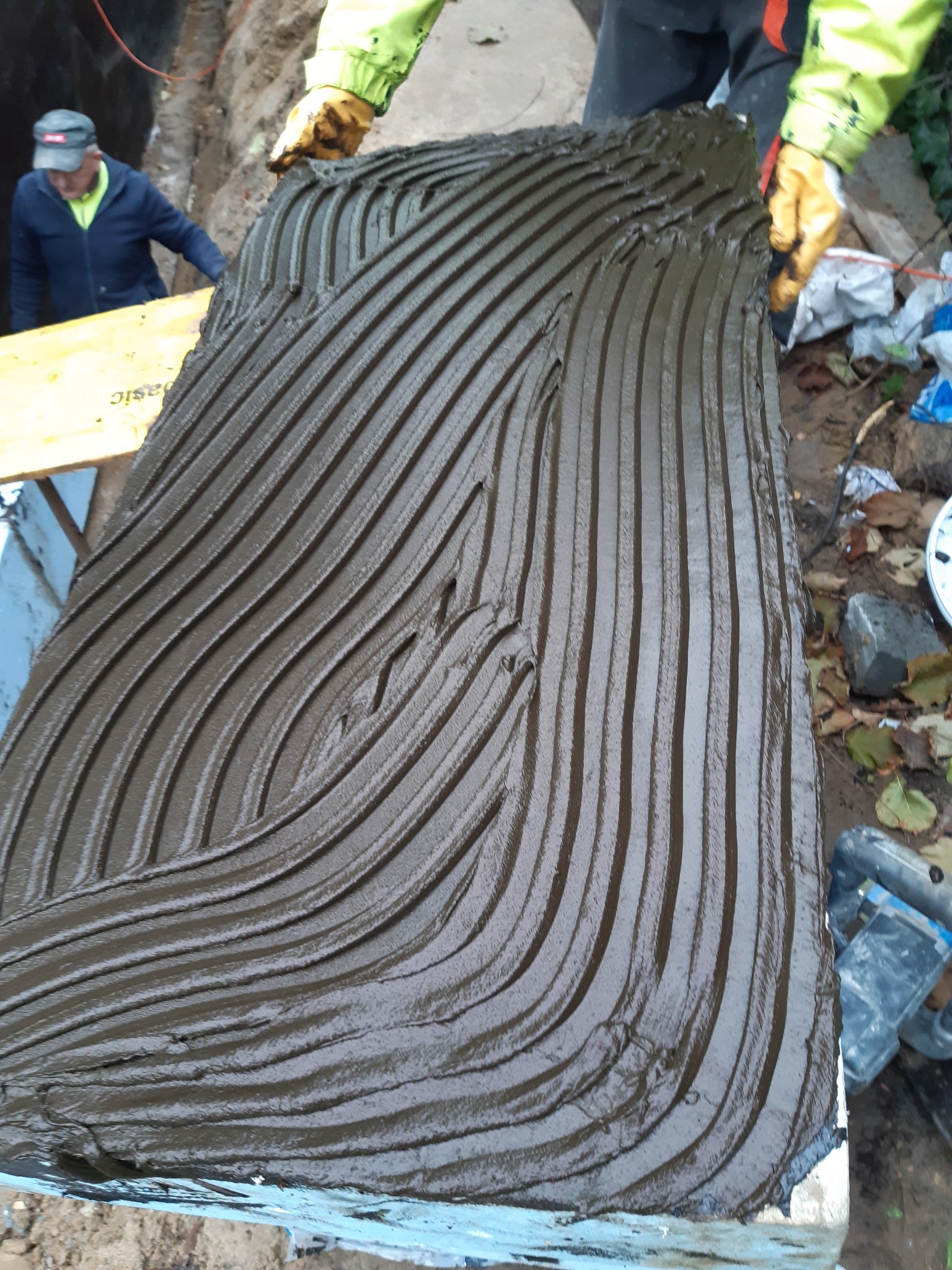 Przygotowanie izolacji fundamentów: nałożona warstwa hydroizolacyjna z charakterystycznymi rowkami, widoczne rękawice robocze i fragment wykopu.