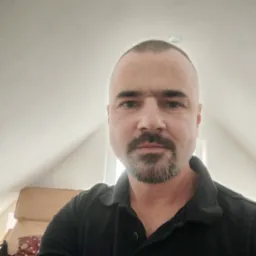 Portret mężczyzny z krótkimi włosami i brodą w czarnej koszulce polo, siedzącego w pokoju na poddaszu, w tle widać fotel.