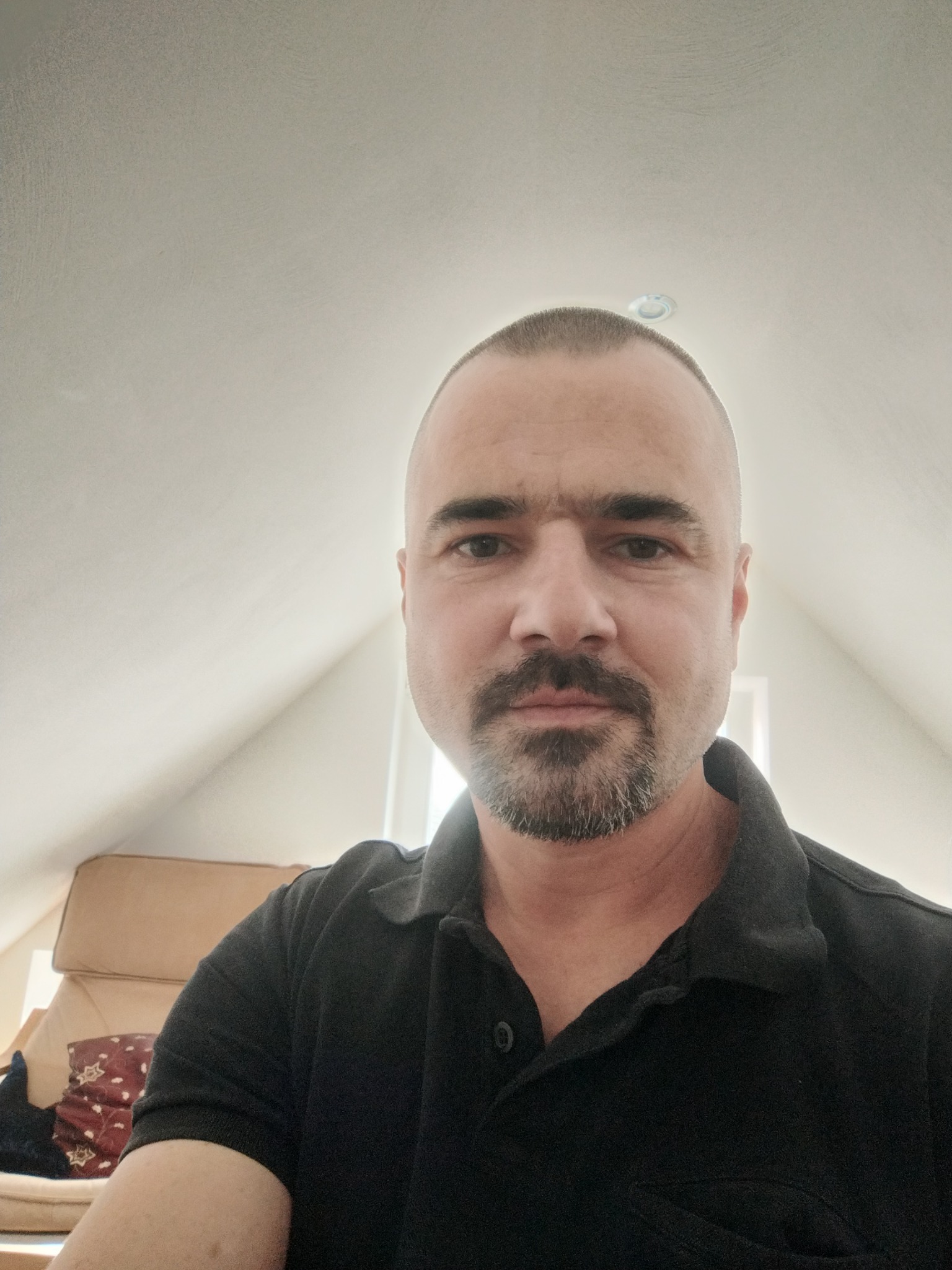 Portret mężczyzny z krótkimi włosami i brodą w czarnej koszulce polo, siedzącego w pokoju na poddaszu, w tle widać fotel.