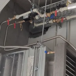 Szara centrala wentylacyjna podwieszona pod sufitem z widocznymi rurami i zaworami. Metalowe elementy konstrukcyjne i instalacyjne.