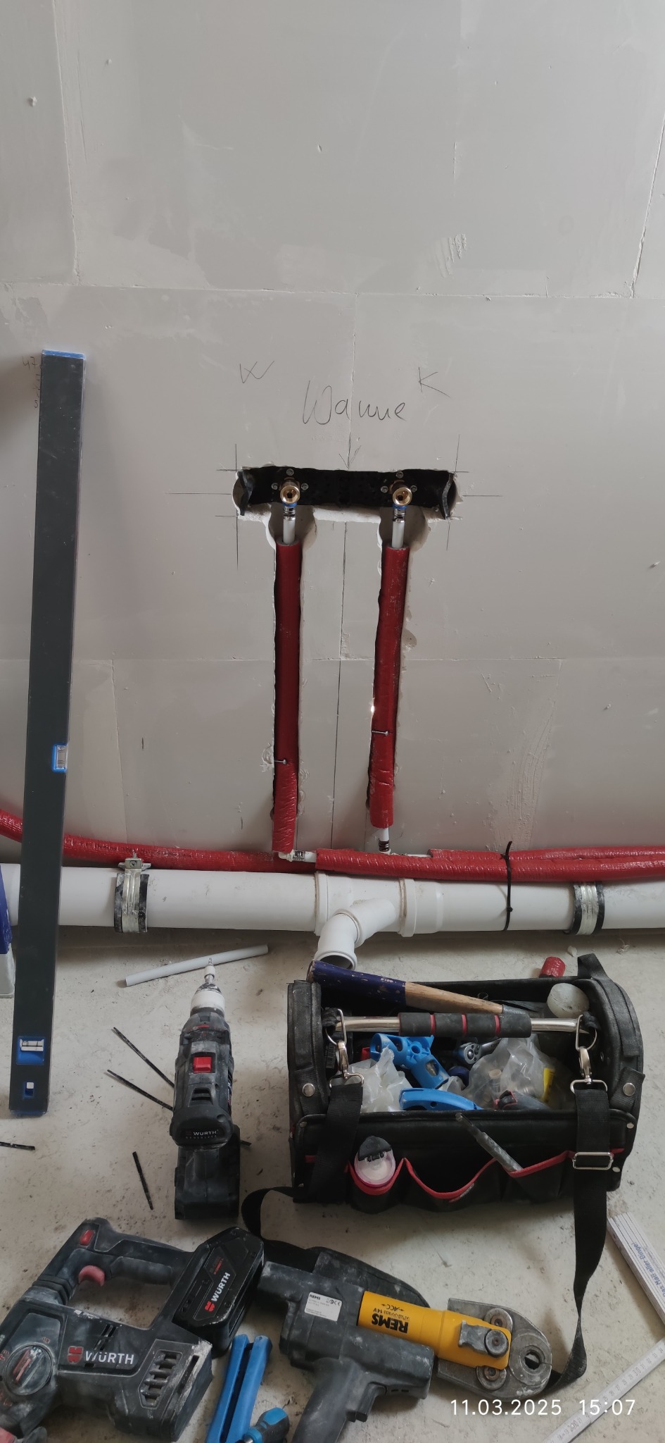 Montaż instalacji wodnej w ścianie z widocznymi rurami w izolacji, narzędziami i skrzynką narzędziową na podłodze. Widoczny napis 'Wanne'.