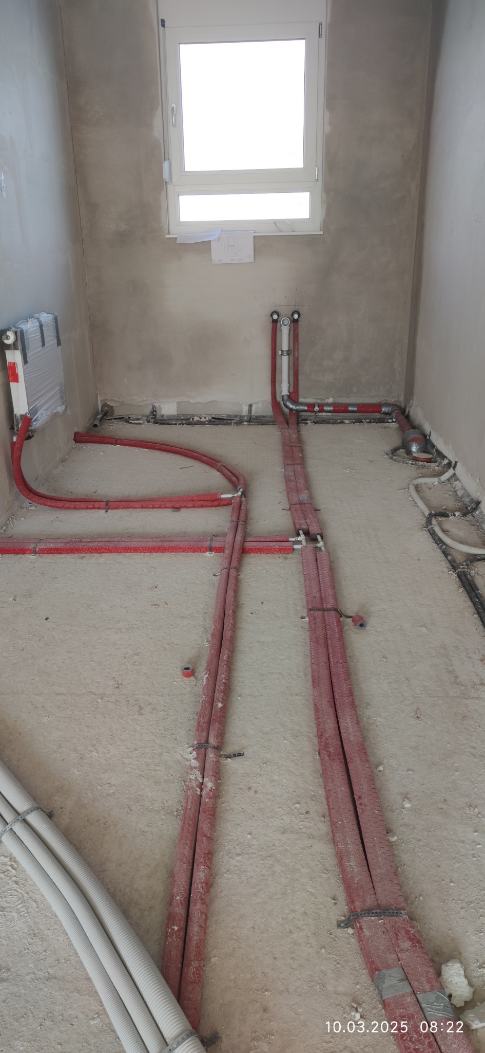Instalacja ogrzewania podłogowego w surowym stanie: czerwone rury w izolacji, rozdzielacz na ścianie, okno w tle. Widok z góry na etapie montażu.