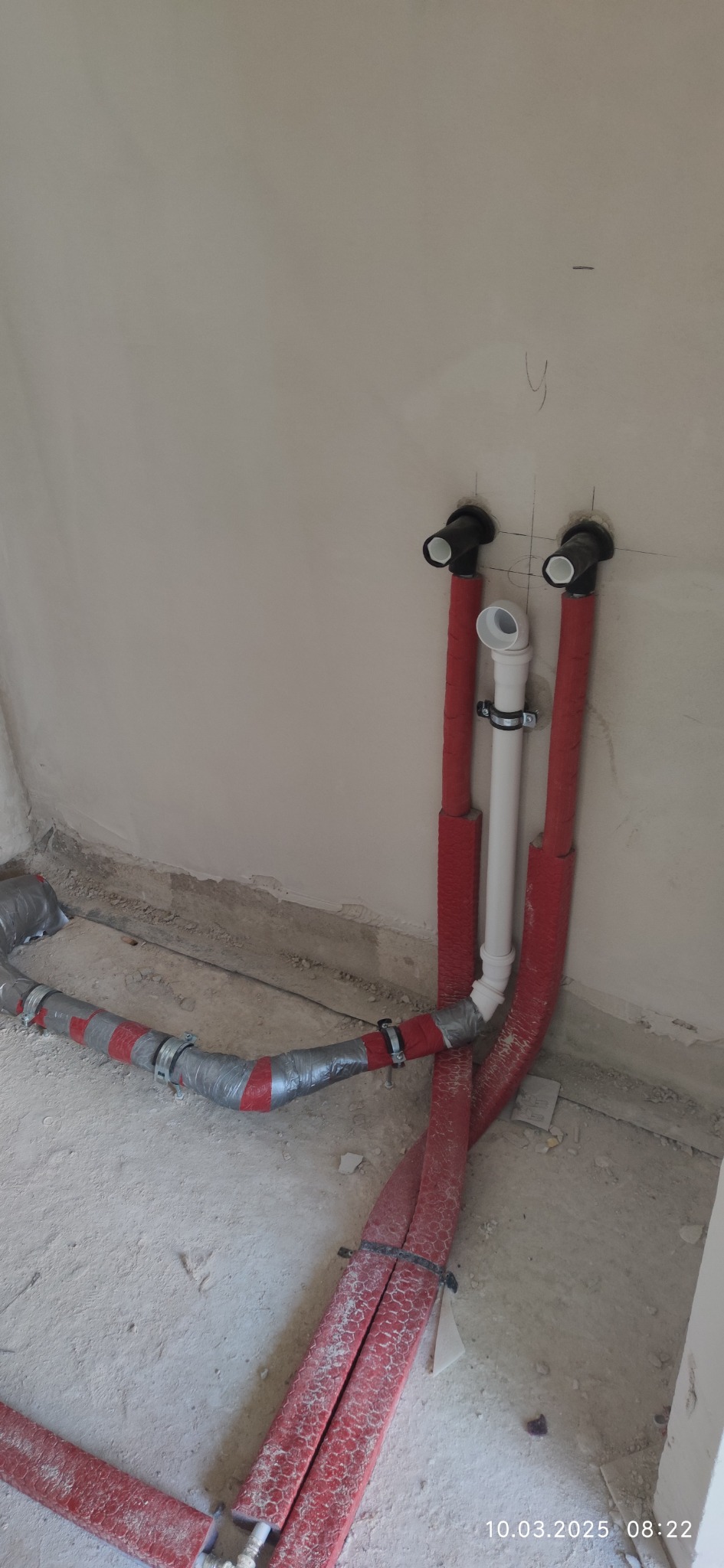 Fragment instalacji hydraulicznej w surowym wnętrzu, z rurami w czerwonej izolacji i elementami montażowymi, widoczne ślady prac budowlanych.