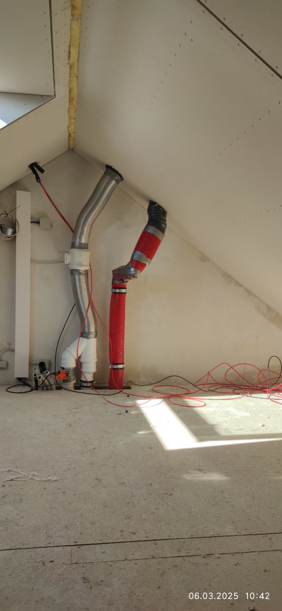 Montaż przewodów wentylacyjnych w przestrzeni poddasza. Widoczne izolowane i metalowe rury, kable oraz elementy mocujące. Prace instalacyjne w trakcie realizacji.