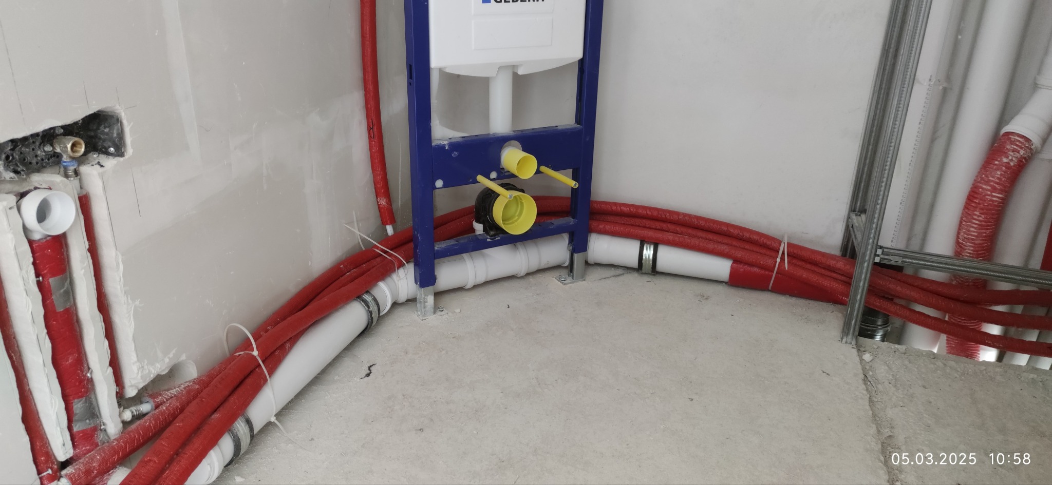 Instalacja podtynkowa Geberit z czerwonymi rurami i białymi odpływami, widoczne mocowania do ściany. Surowy stan, przygotowanie do wykończenia łazienki.
