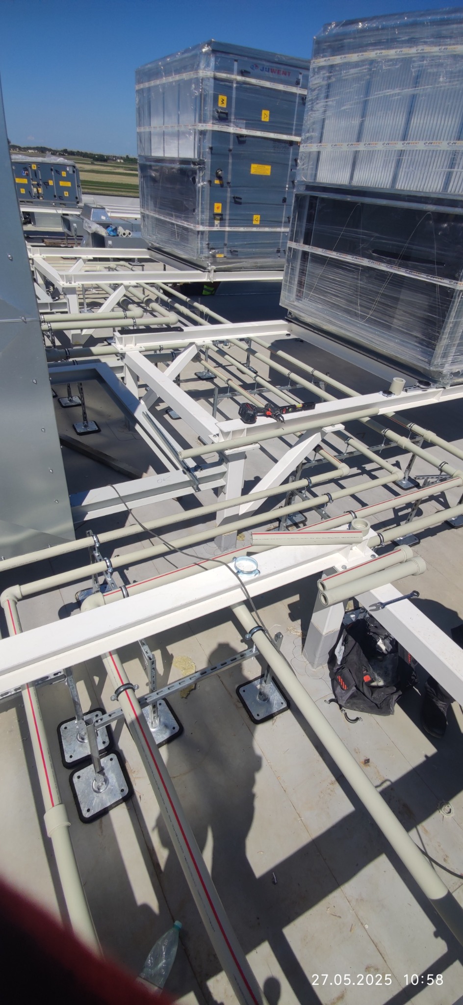 Instalacja wentylacji na dachu: widok z góry na białe rury i srebrne moduły wentylacyjne Juwent, częściowo owinięte folią. Elementy montażowe i narzędzia pracy.