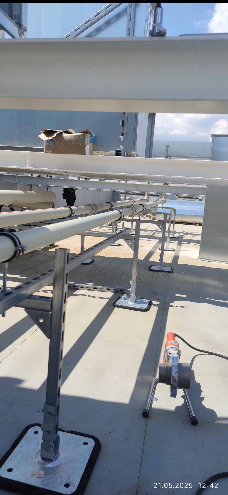 Instalacja rur wentylacyjnych na dachu, widoczne wsporniki, rury z obejmami i zgrzewarka do rur. Częściowo widoczne niebo.