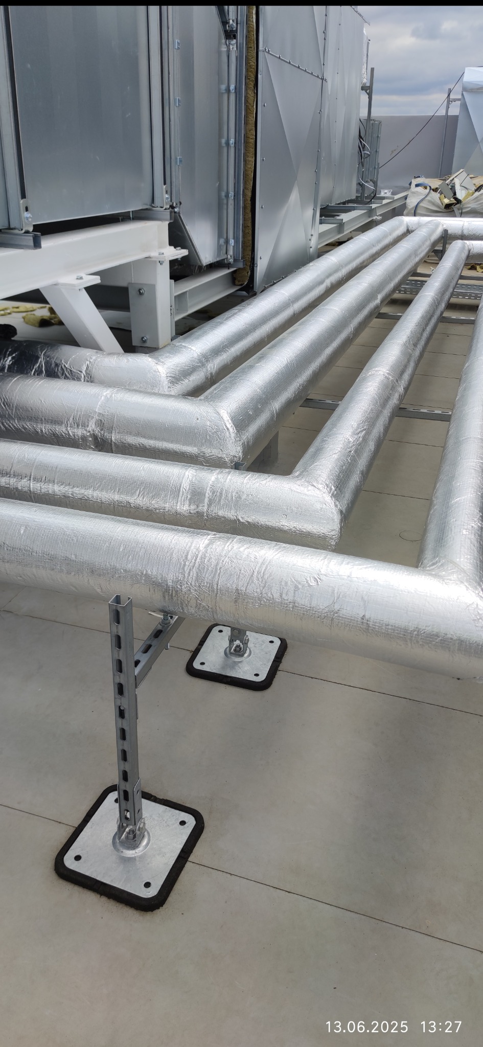 System rur wentylacyjnych w srebrnej izolacji na dachu, wsparty na metalowych konstrukcjach. Widoczne elementy mocujące i fragmenty instalacji na zewnątrz budynku.