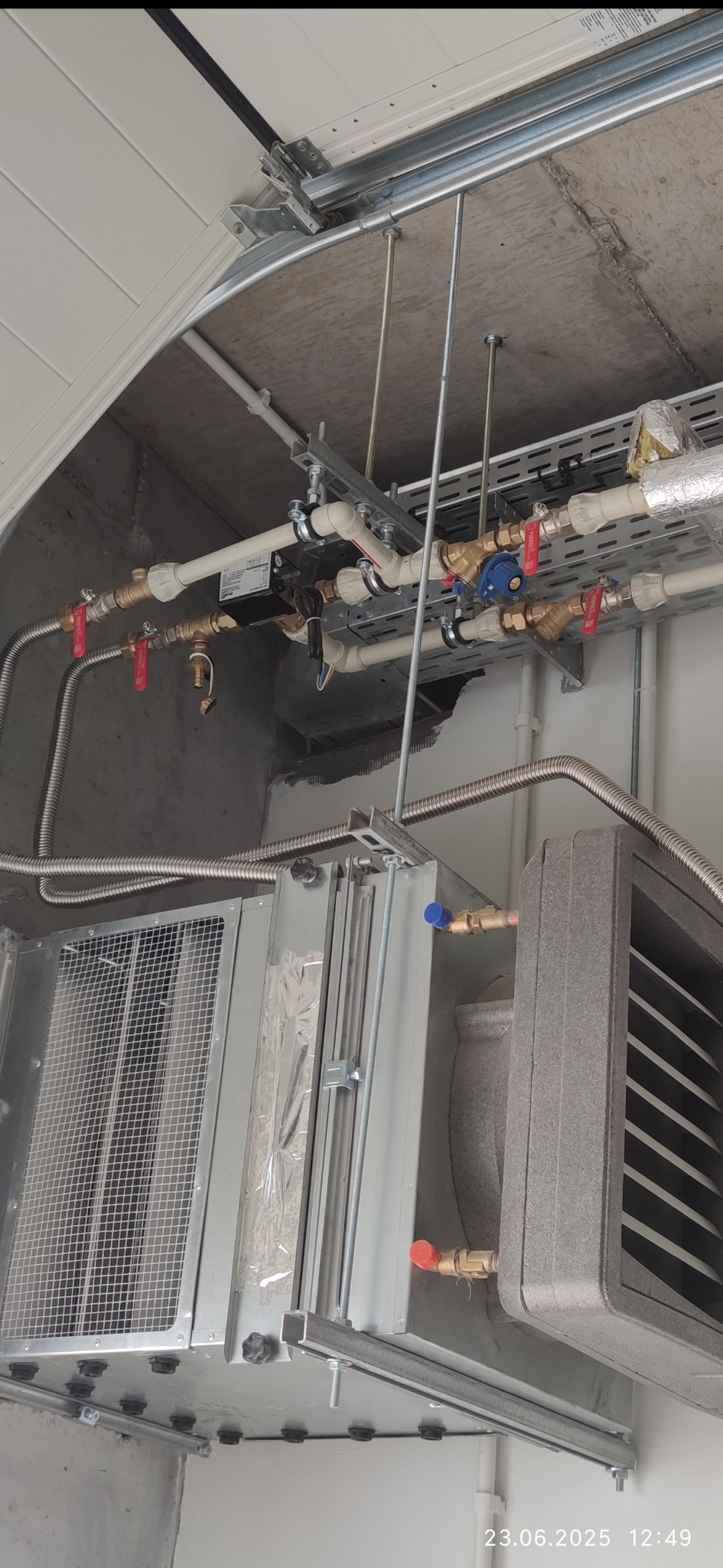 Szara centrala wentylacyjna podwieszona pod sufitem z widocznymi rurami i zaworami. Metalowe elementy konstrukcyjne i instalacyjne.