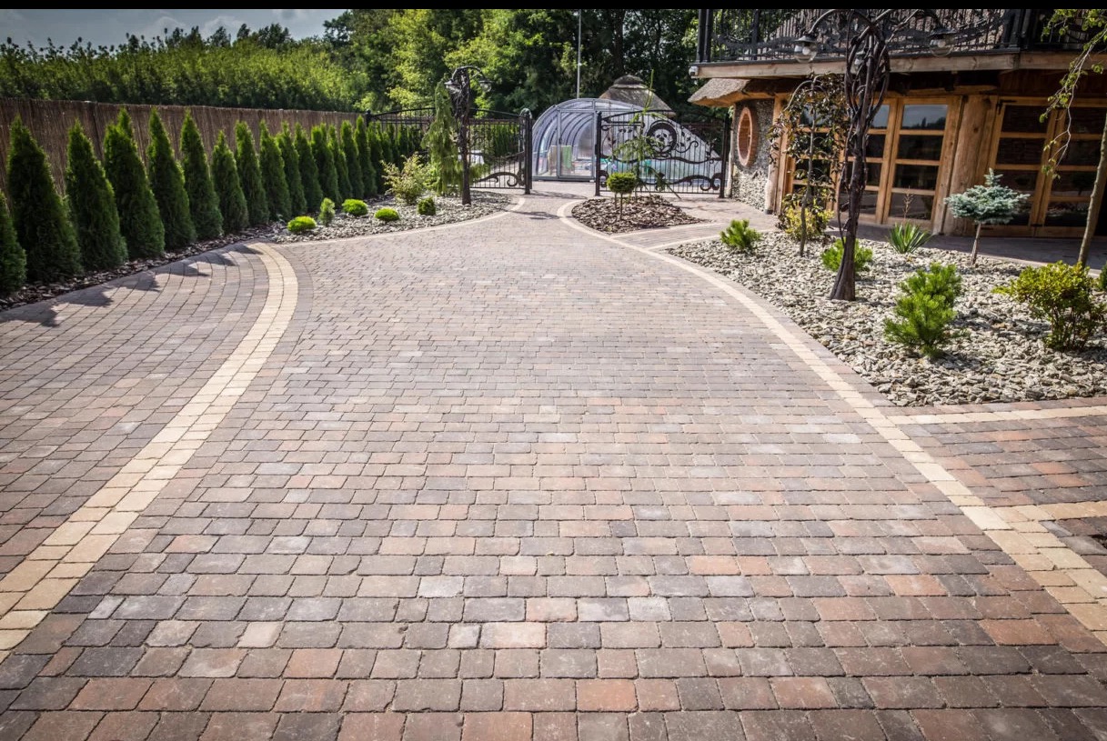 Ułożona z kostki brukowej aleja prowadząca do domu z drewnianą fasadą i ogrodem z iglakami. Jasna fuga podkreśla kształt kostki.