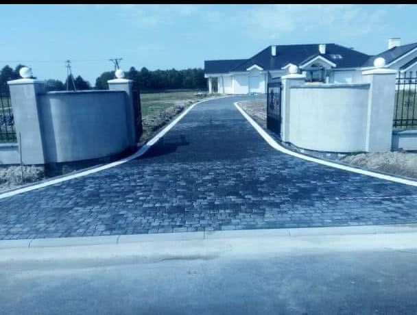 Czarna kostka brukowa ułożona na podjeździe do domu z bramą, obrzeża z białej kostki. W oddali widoczny dom z ciemnym dachem.