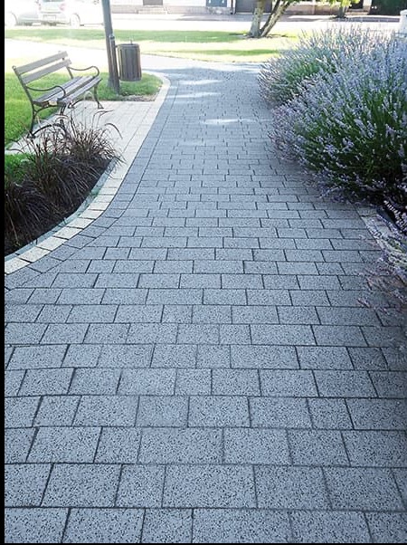 Szary chodnik z kostki brukowej, obrzeże z jasnych kostek, rabaty z lawendą i ozdobnymi trawami, ławka w tle.