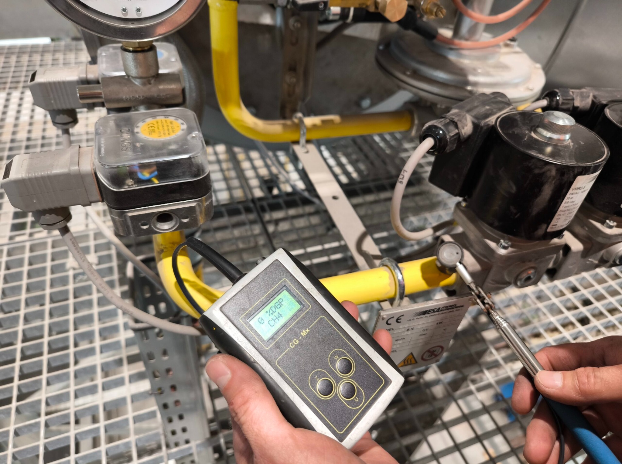 Gazownik wykonuje pomiar szczelności instalacji gazowej za pomocą detektora gazu. Żółte rury instalacji, czujnik w dłoni, metalowa kratka w tle.