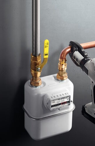 Biały gazomierz z żółtym zaworem i miedzianymi rurami, częściowo widoczne narzędzie zaciskowe po prawej stronie, na tle szarej ściany.