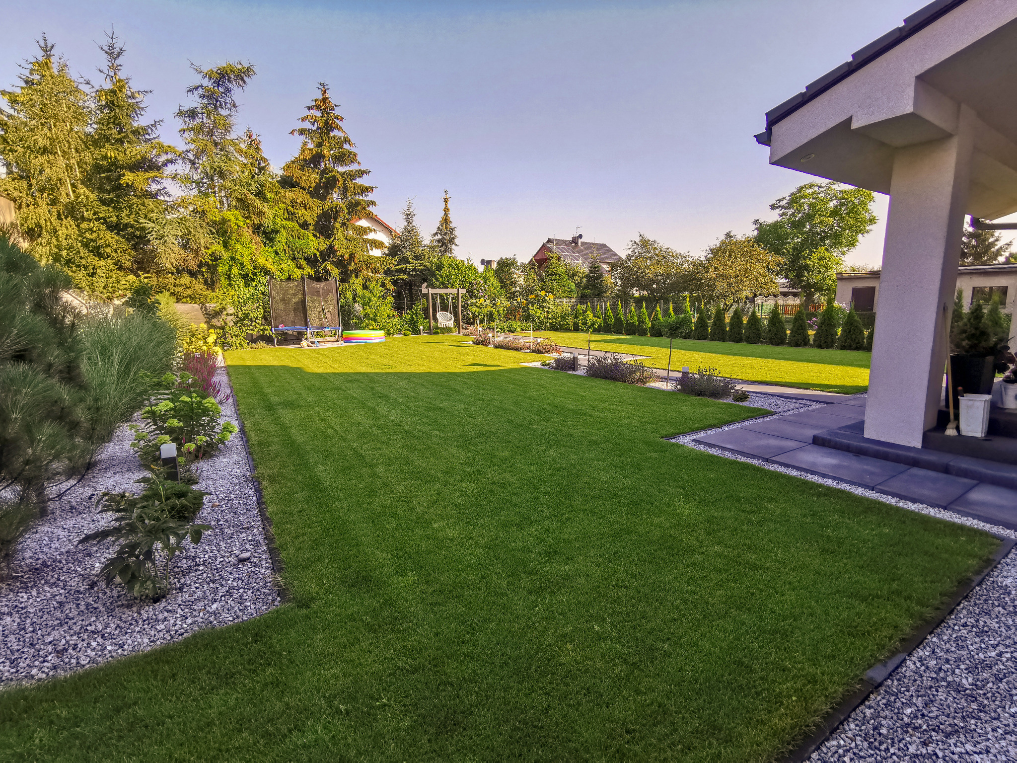 Zadbany trawnik z równo skoszoną trawą, obrzeża z kamienia i roślin, w tle drzewa iglaste i trampolina, widok z perspektywy tarasu nowoczesnego domu.