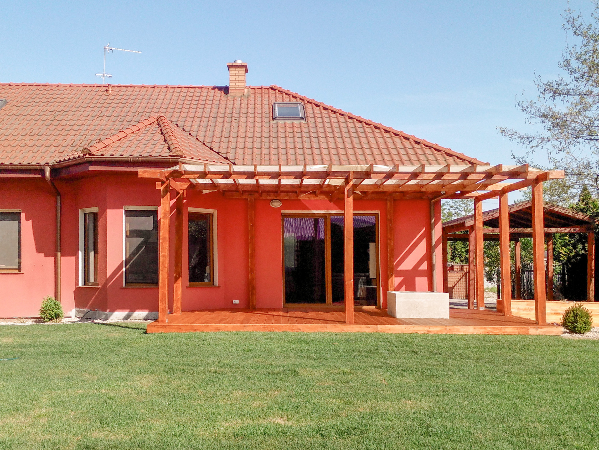 Czerwony dom z drewnianym tarasem i pergolą, zielony trawnik. Widoczne okna, drzwi balkonowe i fragment ogrodu. Słoneczny dzień.