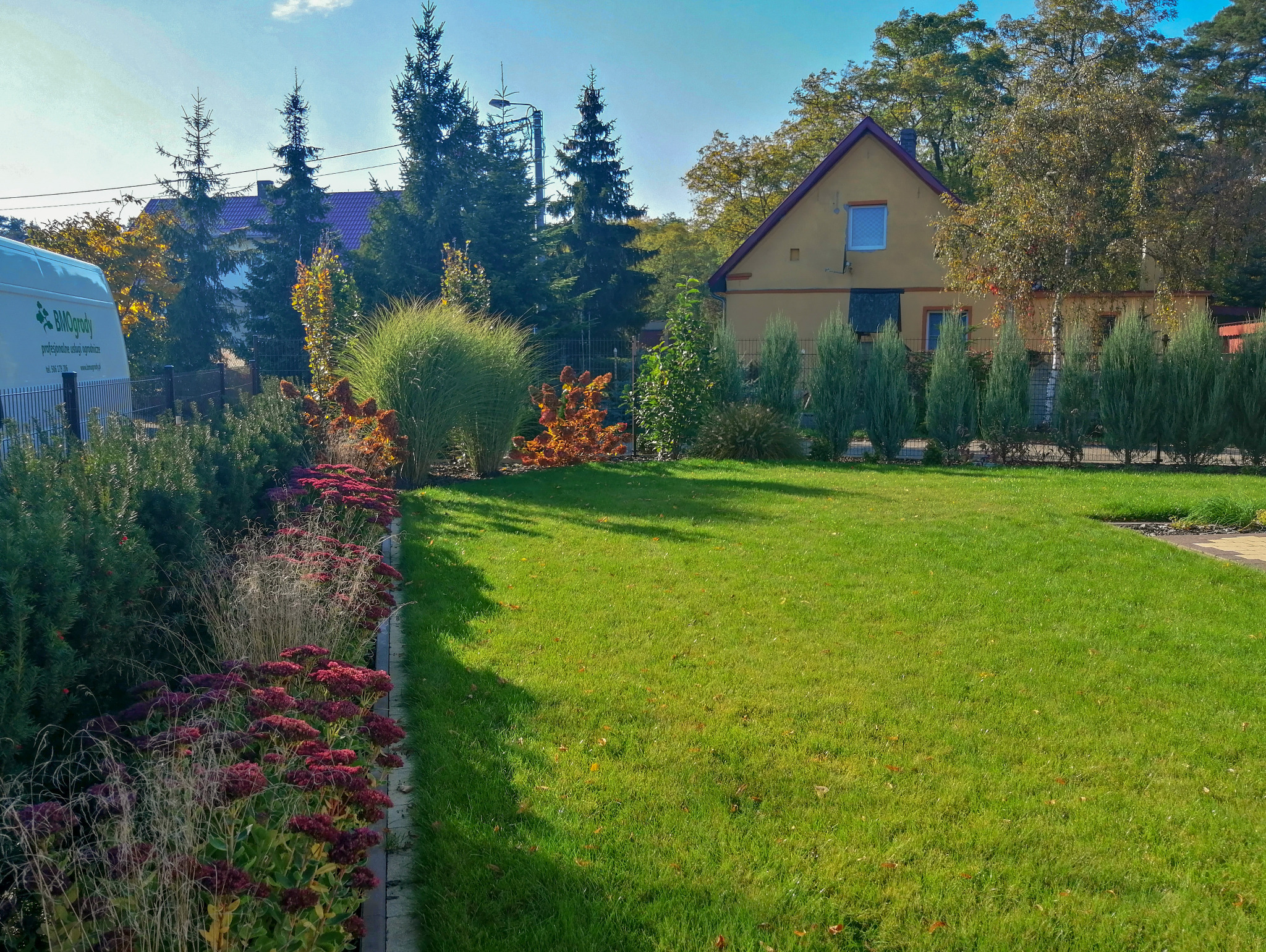 Zadbany trawnik z rabatami kwiatowymi i ozdobnymi krzewami na tle domu z ogrodem. Widoczny fragment firmowego busa.