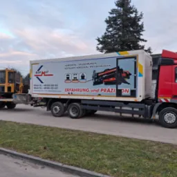 Czerwona ciężarówka z naczepą, na której widnieje grafika urządzenia do przewiertów. Na przyczepie koparka Vermeer. Sprzęt do prac ziemnych, przewiertów.