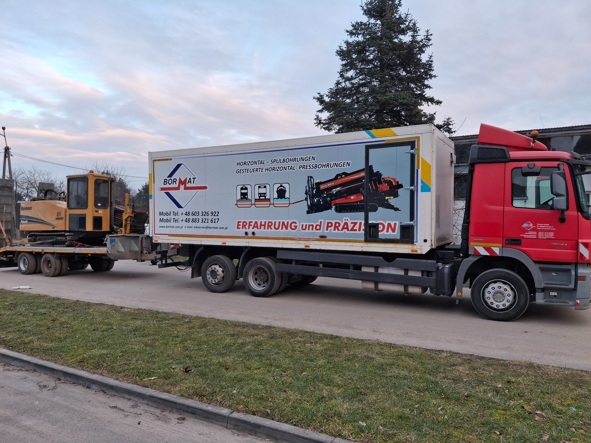 Czerwona ciężarówka z naczepą, na której widnieje grafika urządzenia do przewiertów. Na przyczepie koparka Vermeer. Sprzęt do prac ziemnych, przewiertów.