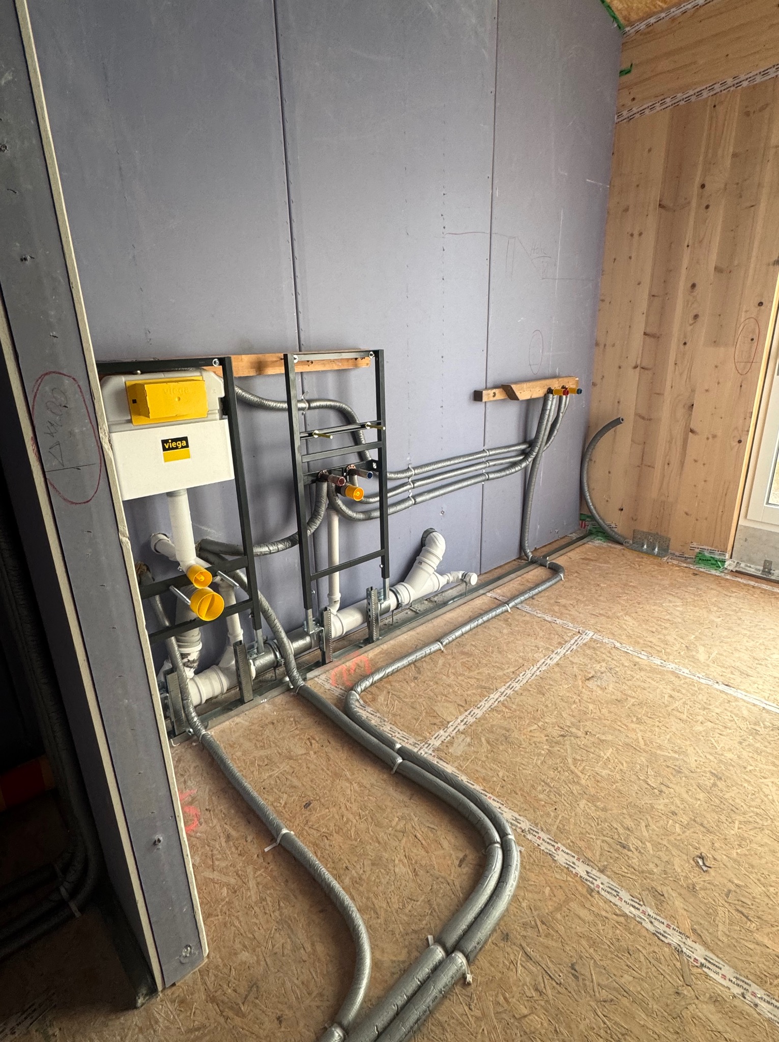 Szara instalacja wodno-kanalizacyjna Viega z żółtymi elementami, zamontowana na ścianie z płyt OSB i szarej płyty gipsowej, widok z góry na podłogę z płyty OSB.