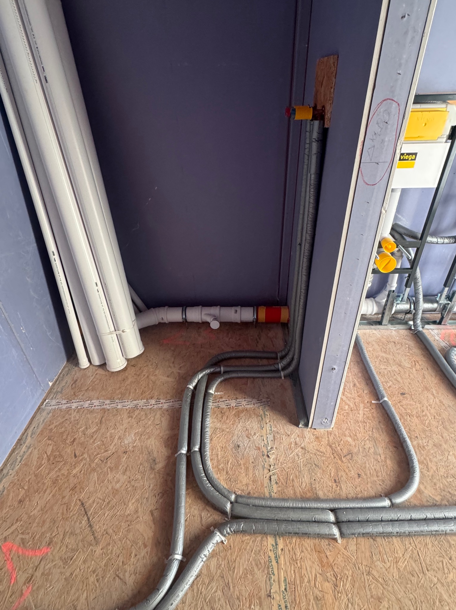 Szare, elastyczne rury instalacji grzewczej ułożone na płycie OSB, z widocznymi białymi rurami kanalizacyjnymi i fragmentem stelaża podtynkowego WC.