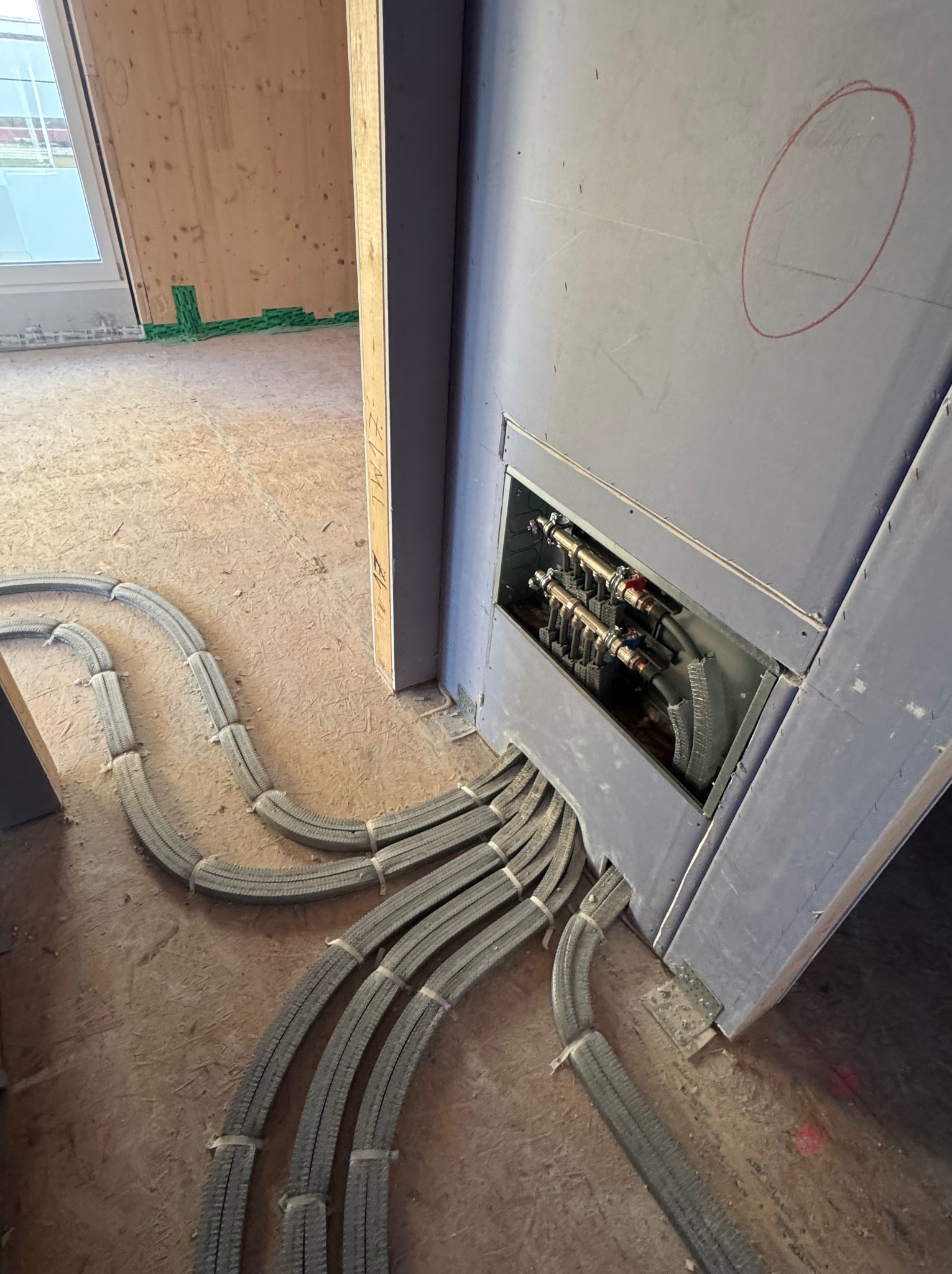 Szare, elastyczne rury instalacji wodnej wchodzące do rozdzielacza w ścianie z płyty gipsowej na surowej podłodze. Widok z góry, w tle okno i drewniana ściana.