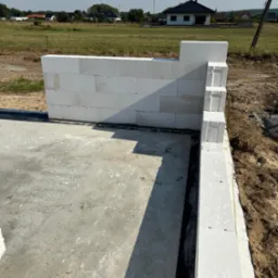 Wznoszenie ścian fundamentowych z białych bloczków na płycie fundamentowej. Widok na budowę domu jednorodzinnego w otoczeniu łąk i zabudowań.