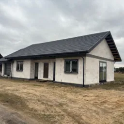 Niedokończony, parterowy dom z białego betonu komórkowego, z czarną dachówką, czarnymi oknami i drzwiami. Widoczne worki z materiałami budowlanymi.