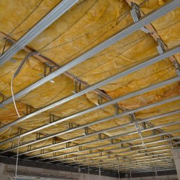 Solidhouse - Montaż izolacji z wełny mineralnej i stelaża sufitu podwieszanego w surowym wnętrzu. Widoczne profile metalowe, drewniane legary i przewody instalacyjne.