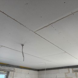 Solidhouse - Sufit z płyt kartonowo-gipsowych, widoczne łączenia i wkręty. W tle surowe ściany z betonu komórkowego i okno. Wystające kable elektryczne.