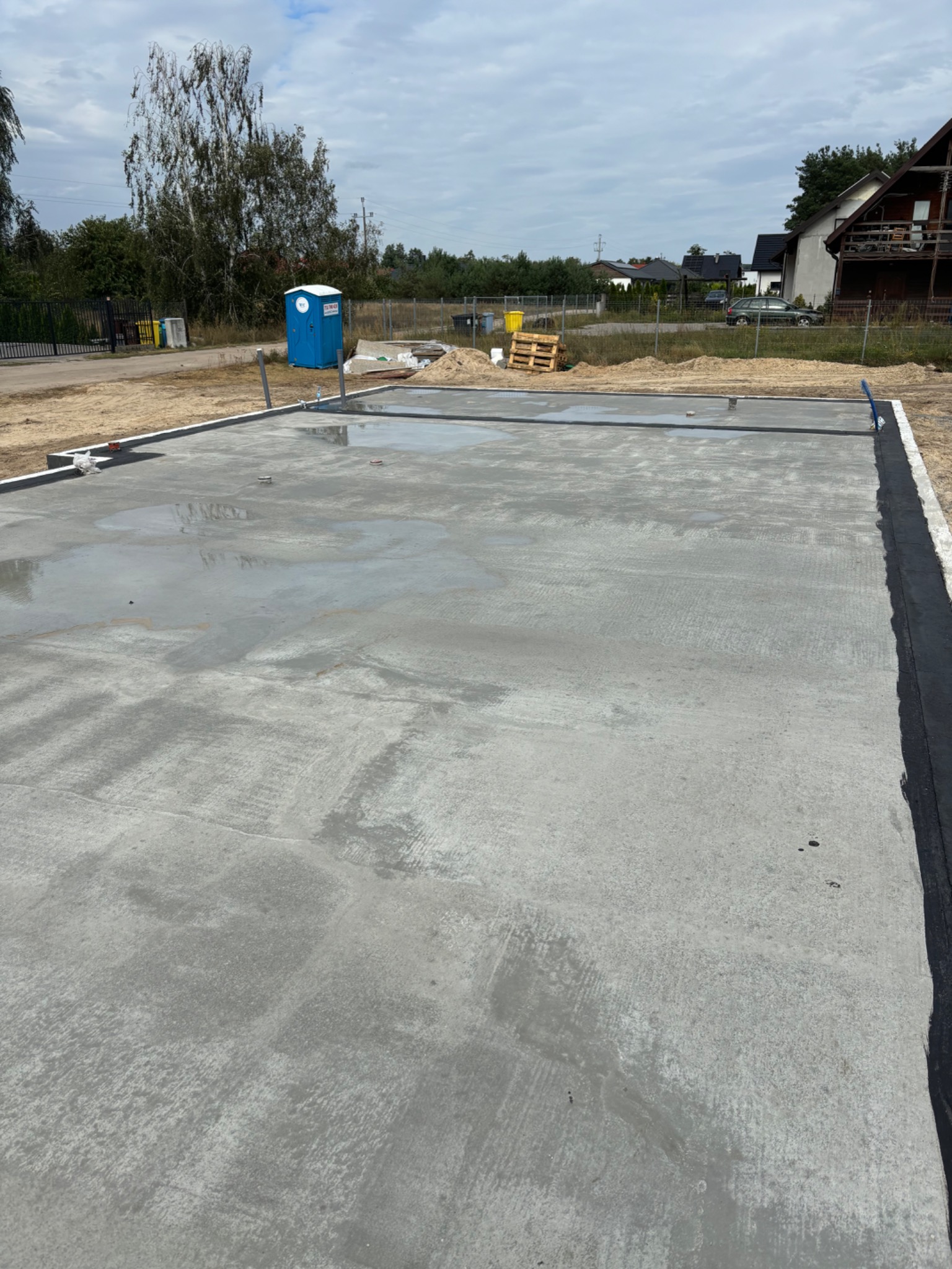 Wylana, świeża płyta fundamentowa na placu budowy z widocznymi elementami szalunku i izolacji. W tle toaleta TOI-TOI, drzewa i zabudowa jednorodzinna.