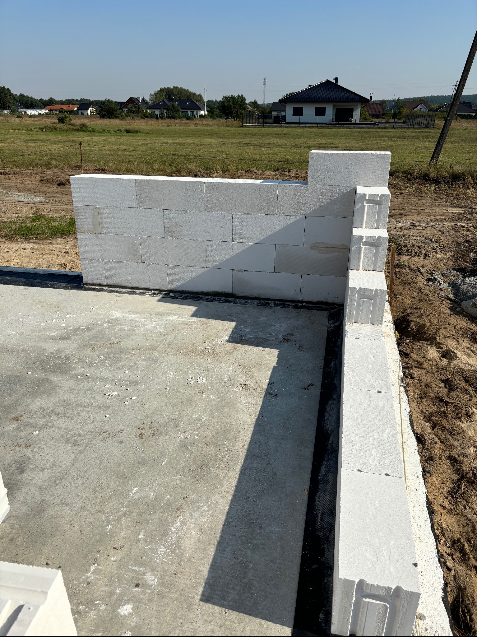 Wznoszenie ścian fundamentowych z białych bloczków na płycie fundamentowej. Widok na budowę domu jednorodzinnego w otoczeniu łąk i zabudowań.