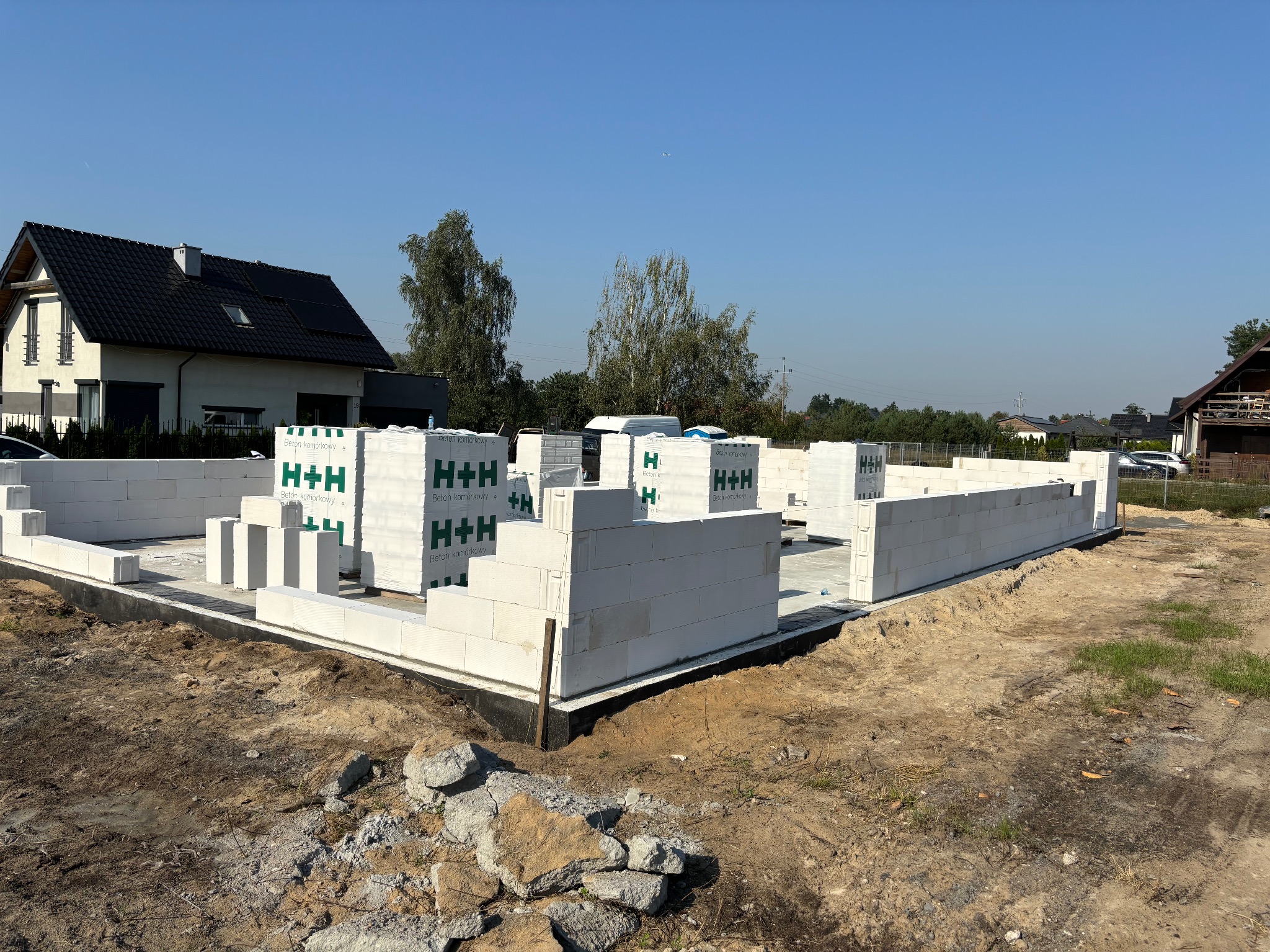 Budowa domu z betonu komórkowego H+H: wzniesione ściany fundamentowe na tle zieleni i błękitnego nieba. Widoczne palety z bloczkami.