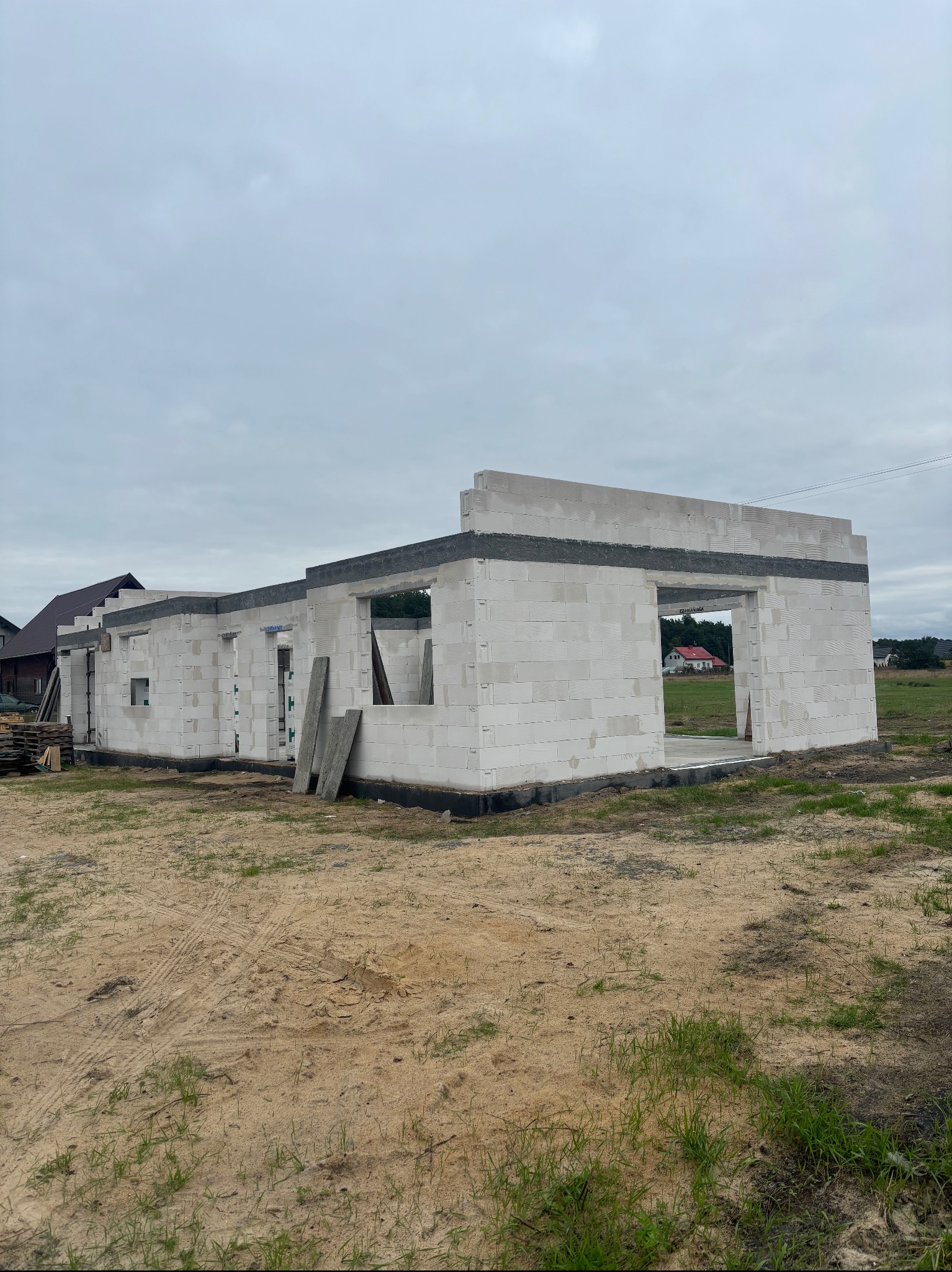 Surowy stan domu z białych bloczków betonu komórkowego. Budynek w trakcie budowy, widoczne otwory okienne i drzwiowe. Na pierwszym planie teren budowy z piaskiem i trawą.