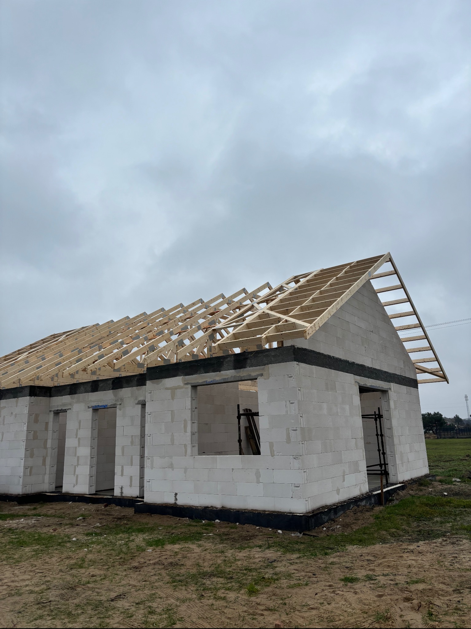 Surowy stan domu z białych bloczków, z widoczną drewnianą konstrukcją dachu i rusztowaniem przy oknie. Tło: pochmurne niebo.