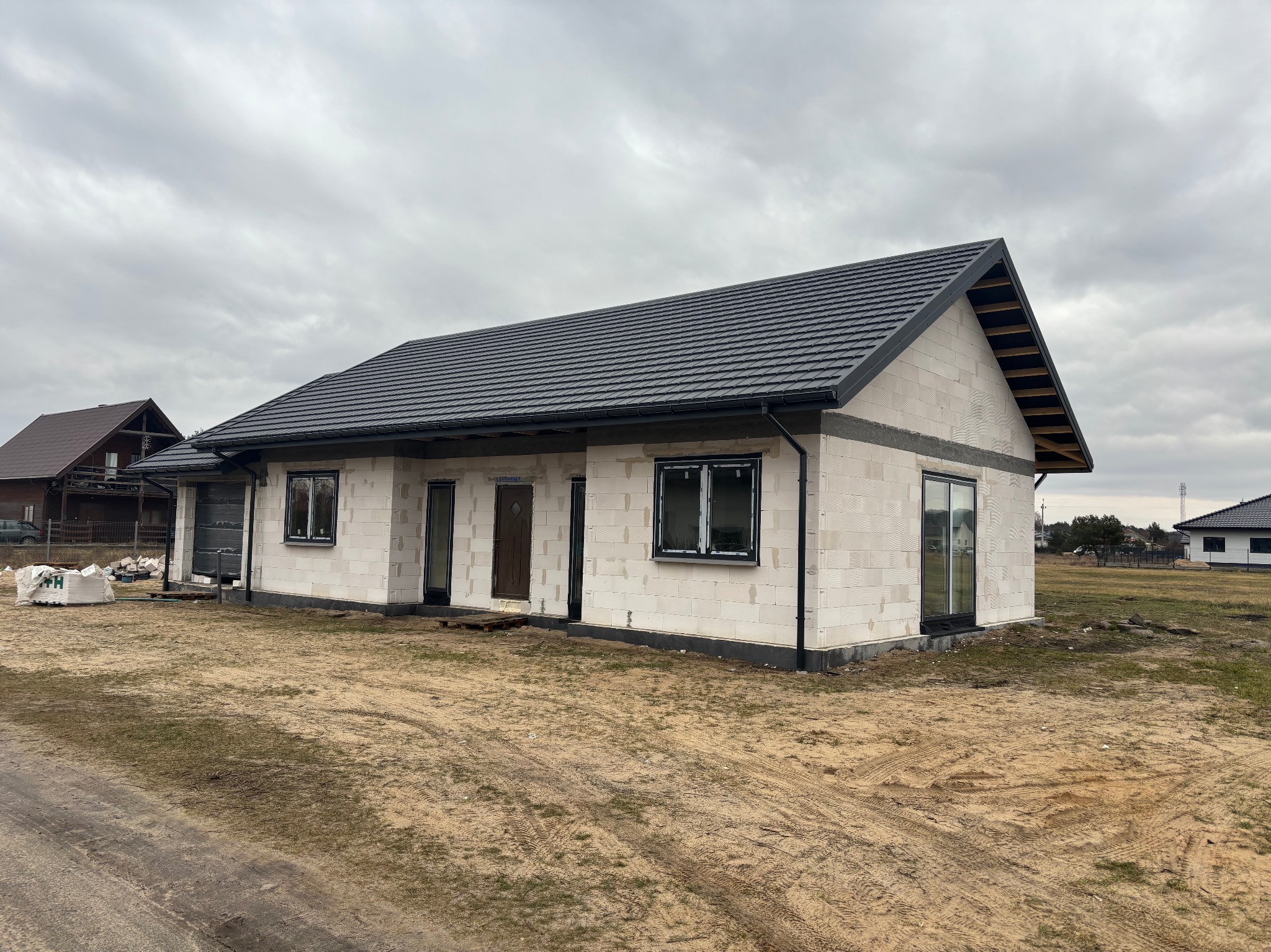 Niedokończony, parterowy dom z białego betonu komórkowego, z czarną dachówką, czarnymi oknami i drzwiami. Widoczne worki z materiałami budowlanymi.
