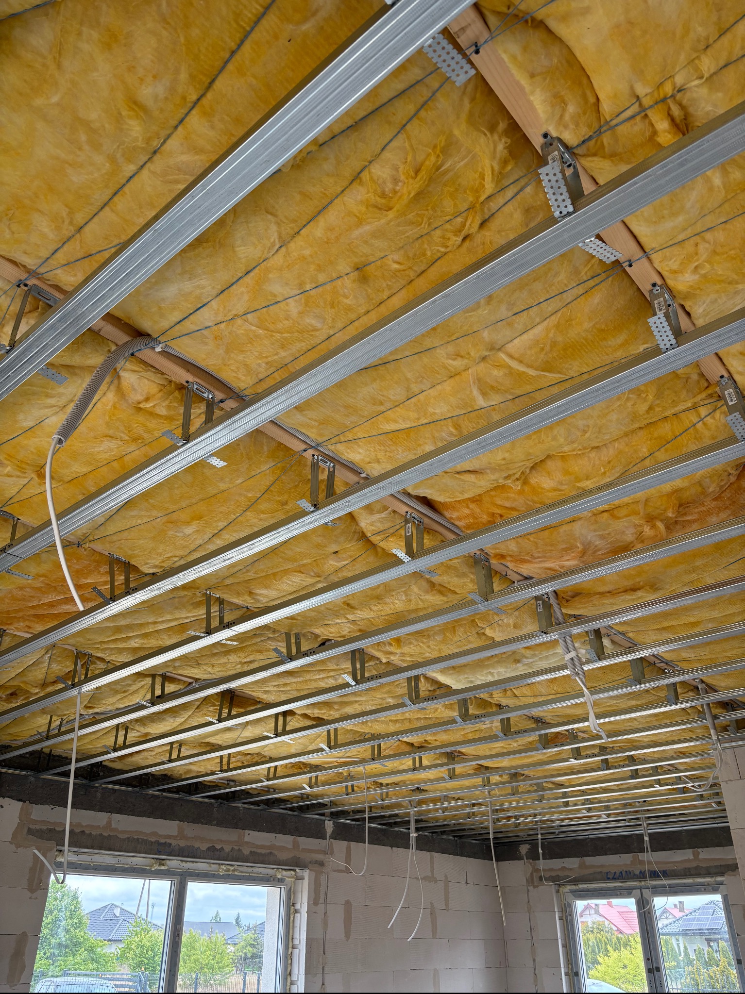 Montaż izolacji z wełny mineralnej i stelaża sufitu podwieszanego w surowym wnętrzu. Widoczne profile metalowe, drewniane legary i przewody instalacyjne.