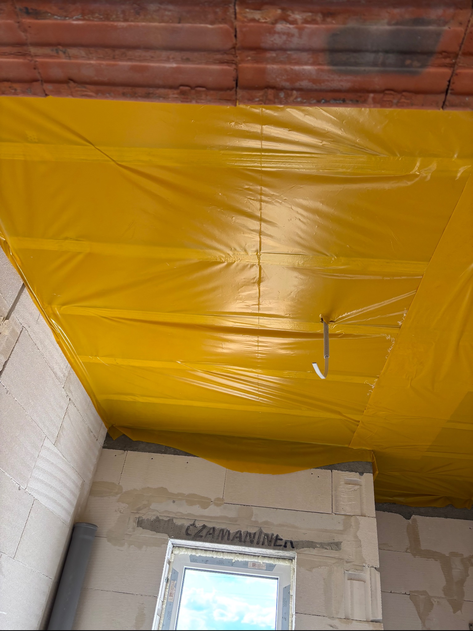 Żółta folia paroizolacyjna szczelnie pokrywa sufit z cegły i ściany z betonu komórkowego, widoczny fragment okna i rury PCV, etap prac budowlanych.