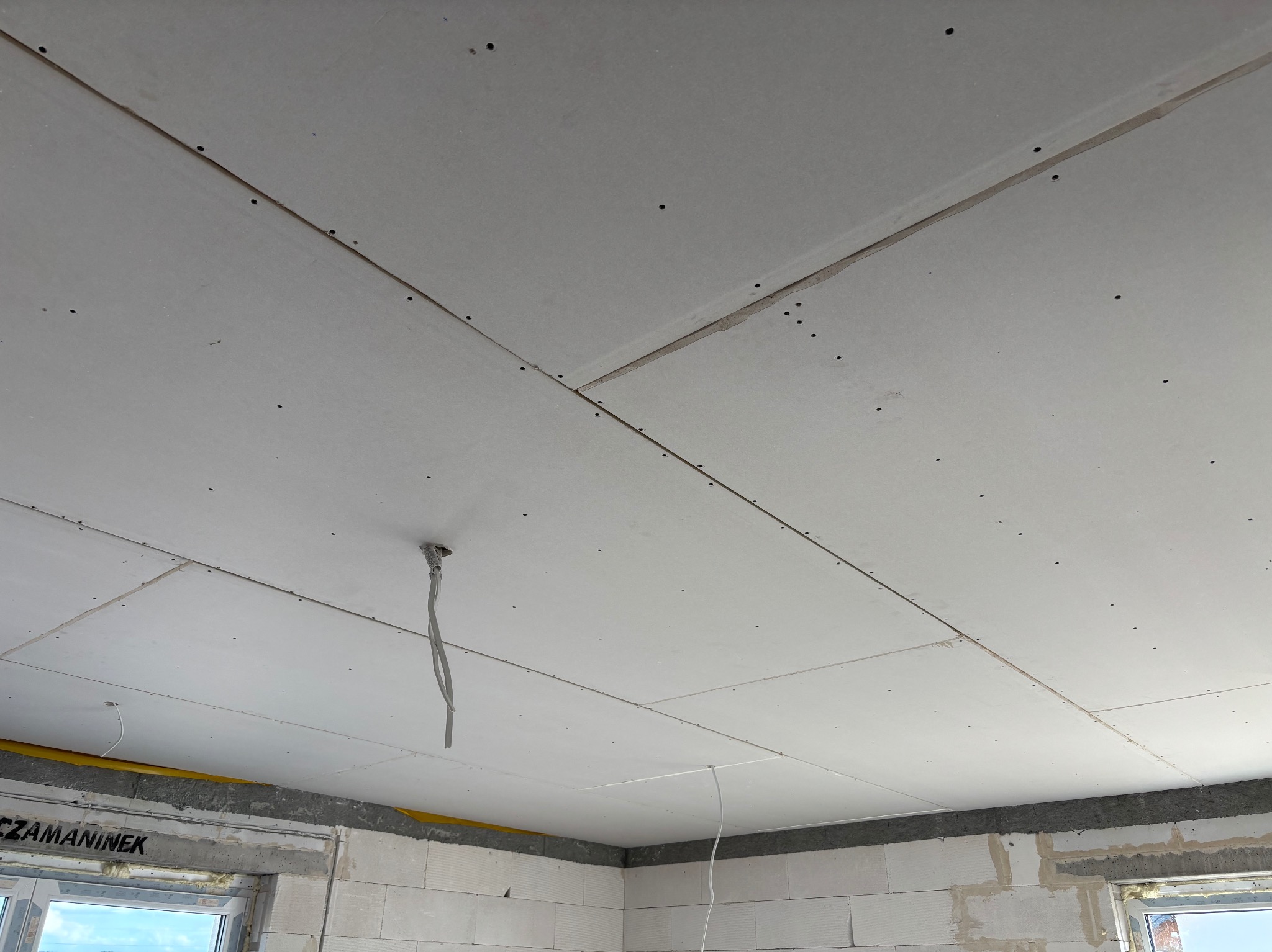 Sufit z płyt kartonowo-gipsowych, widoczne łączenia i wkręty. W tle surowe ściany z betonu komórkowego i okno. Wystające kable elektryczne.