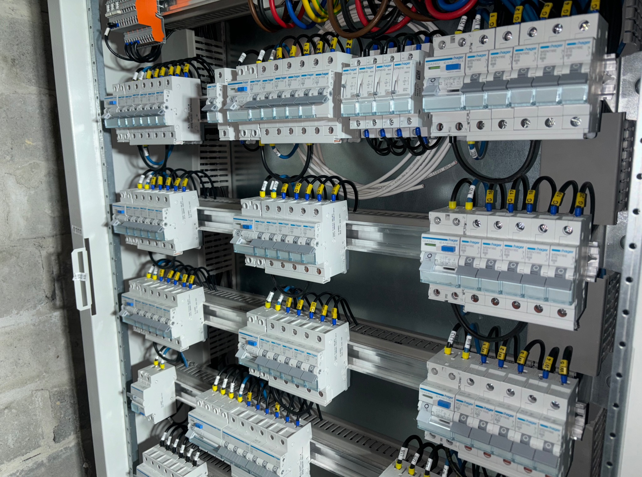 Szczegółowe ujęcie rozdzielnicy elektrycznej z licznymi bezpiecznikami Hager, okablowaniem w kolorach i szarym tłem ściany. Widoczne etykiety na przewodach.