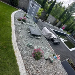 Nowoczesny ogród z basenem, dekoracyjnym żwirem, kamieniami i roślinami. Widoczne drewniane deski tarasu i ozdobne elementy, takie jak wiatraczek i oświetlenie.