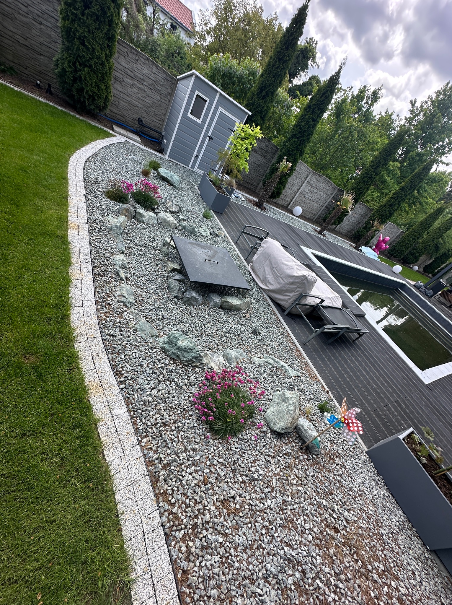 Nowoczesny ogród z basenem, dekoracyjnym żwirem, kamieniami i roślinami. Widoczne drewniane deski tarasu i ozdobne elementy, takie jak wiatraczek i oświetlenie.