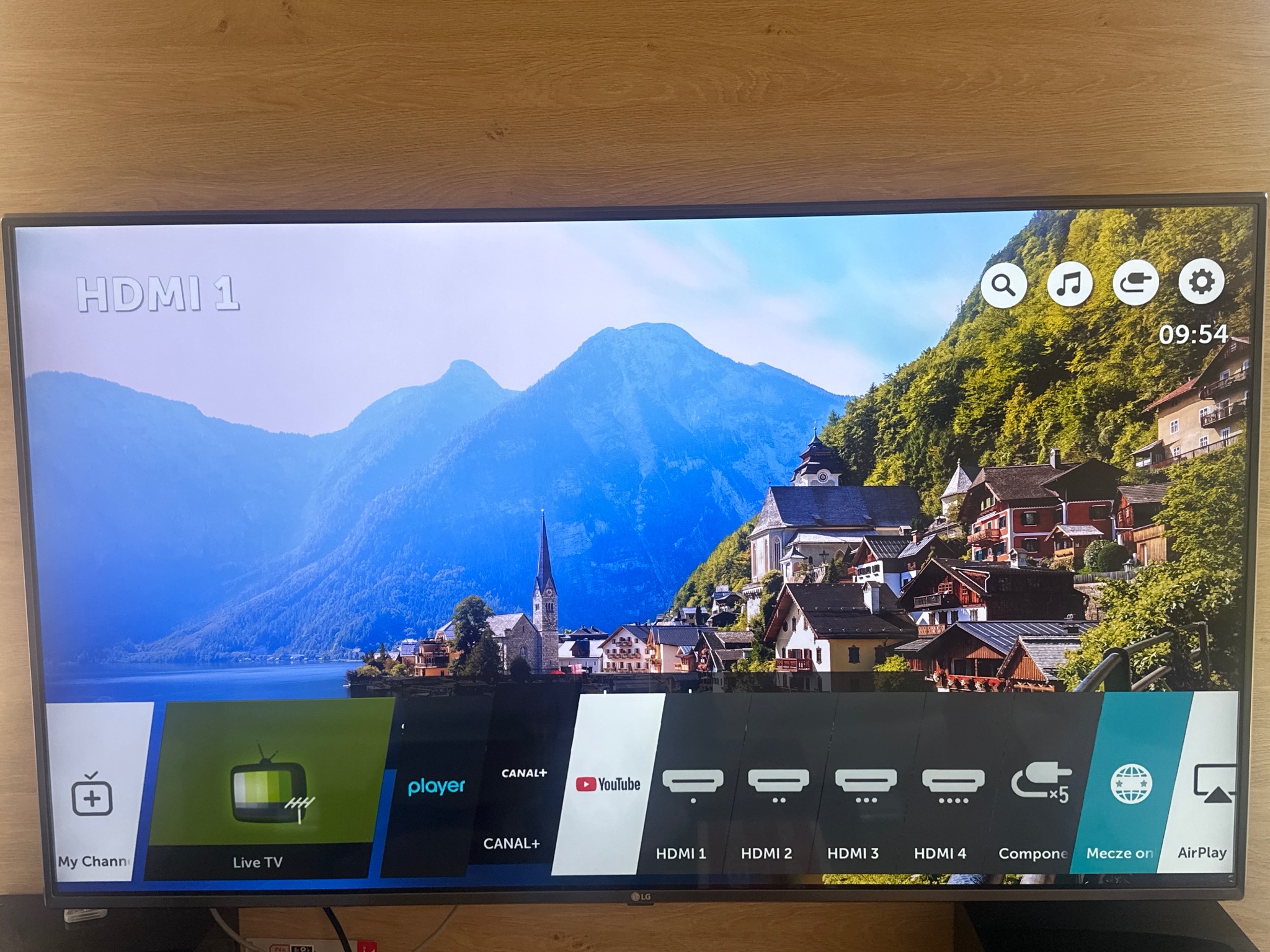 Włączony telewizor LG z widocznym menu wyboru źródła sygnału (HDMI, Live TV, YouTube) i aplikacjami. Na ekranie wyświetlany malowniczy krajobraz górski z jeziorem.