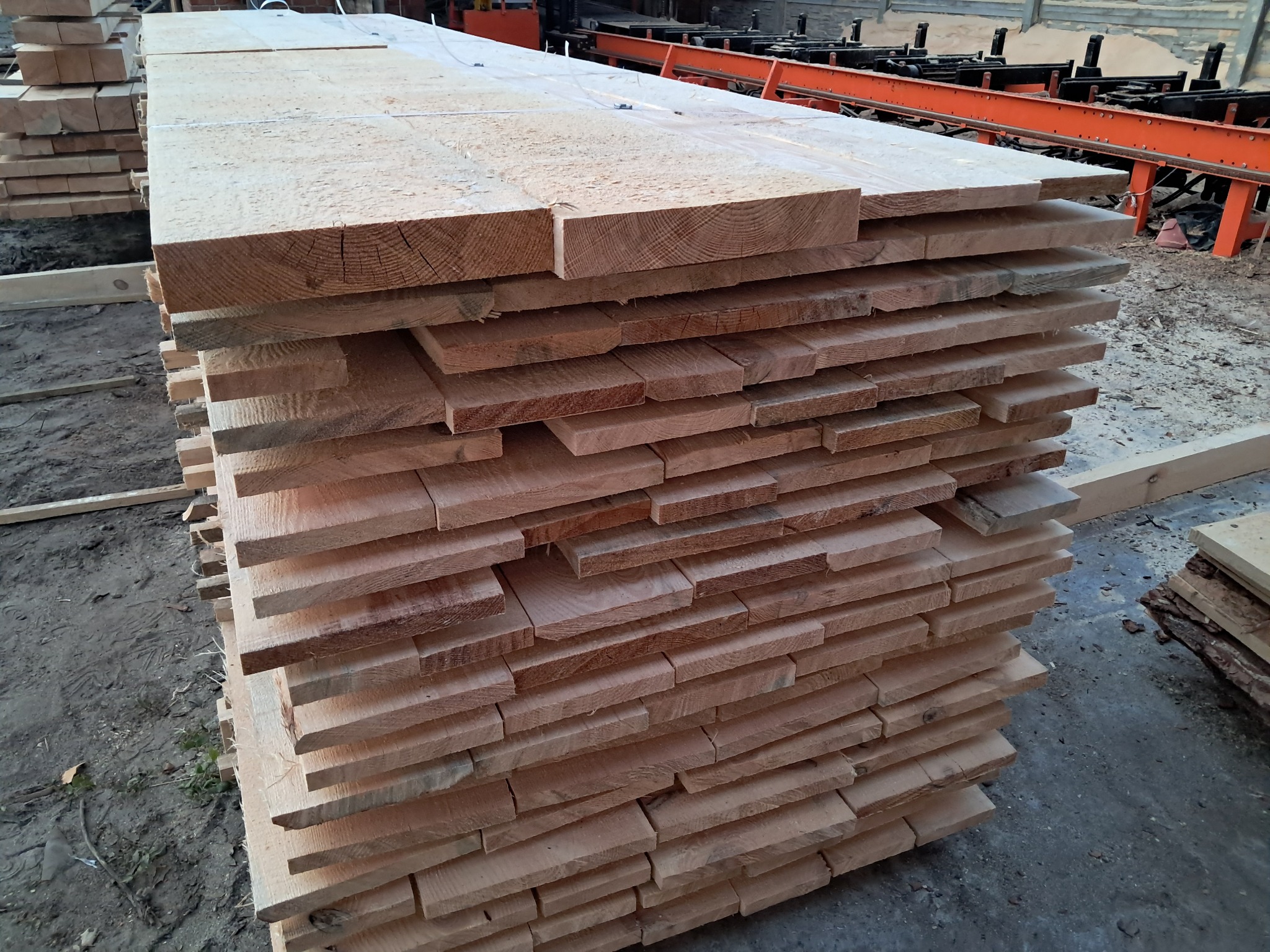 Stos desek z jasnego drewna ułożonych warstwami, widoczne słoje i struktura drewna, w tle pomarańczowa konstrukcja tartaku.