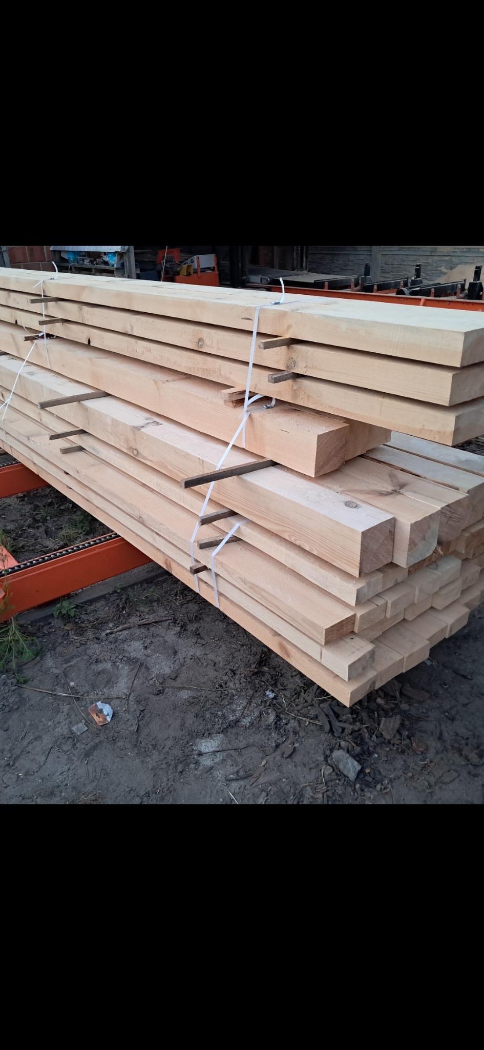 Stos belek drewnianych, spiętych białym sznurkiem, leżących na pomarańczowym regale w tartaku. Widoczne ślady obróbki drewna.