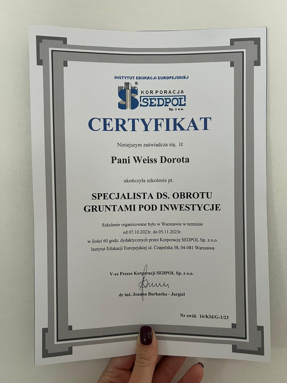 Certyfikat ukończenia szkolenia specjalisty ds. obrotu gruntami pod inwestycje dla Pani Weiss Doroty, wydany przez Korporację SEDPOL. Dokument w szarej ramce trzymany w dłoni.