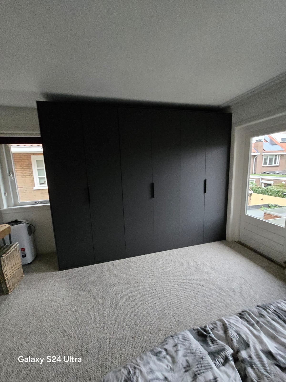 Nowoczesna, czarna szafa wnękowa z minimalistycznymi uchwytami, zamontowana w jasnym pokoju z oknami po obu stronach. Widok na wnętrze pokoju.