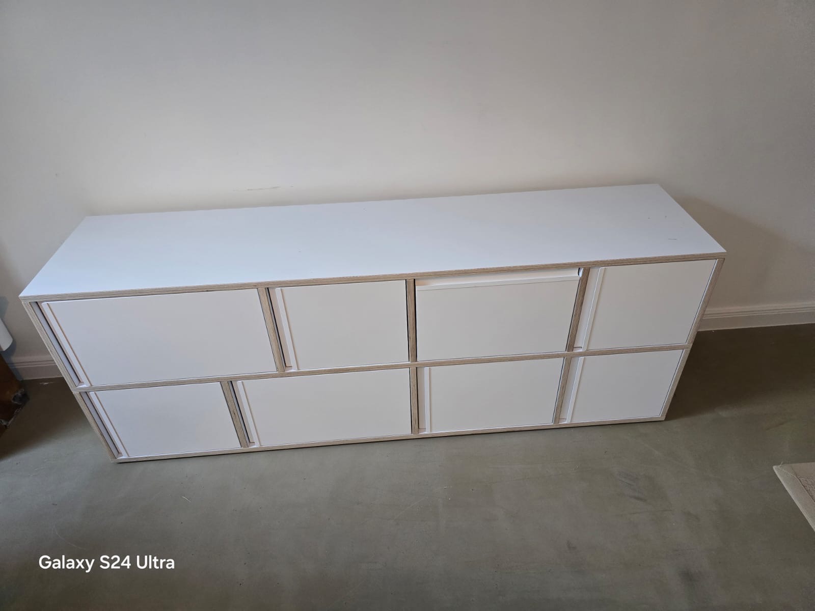 Biała komoda z szufladami, widok z góry, minimalistyczny design, jasne tło. Prosta forma, widoczne krawędzie sklejki, nowoczesny styl.