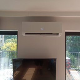 Serhii Cholavyn - Biały klimatyzator ścienny z wyświetlaczem temperatury 24°C, zamontowany w jasnym pokoju z oknami balkonowymi i telewizorem. Widok na zieleń za oknem.