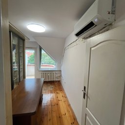 Serhii Cholavyn - Biały klimatyzator na ścianie w pokoju na poddaszu, widoczna instalacja i uszczelnienie pianką montażową. Drewniana podłoga i okno balkonowe w tle.